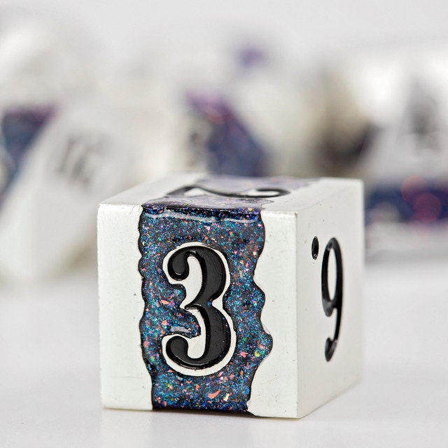 Closeup of a d6 with other metal polyhedral dnd dice in pearl silver metal with beautiful stripes of glittery blue and pink resin in the background.